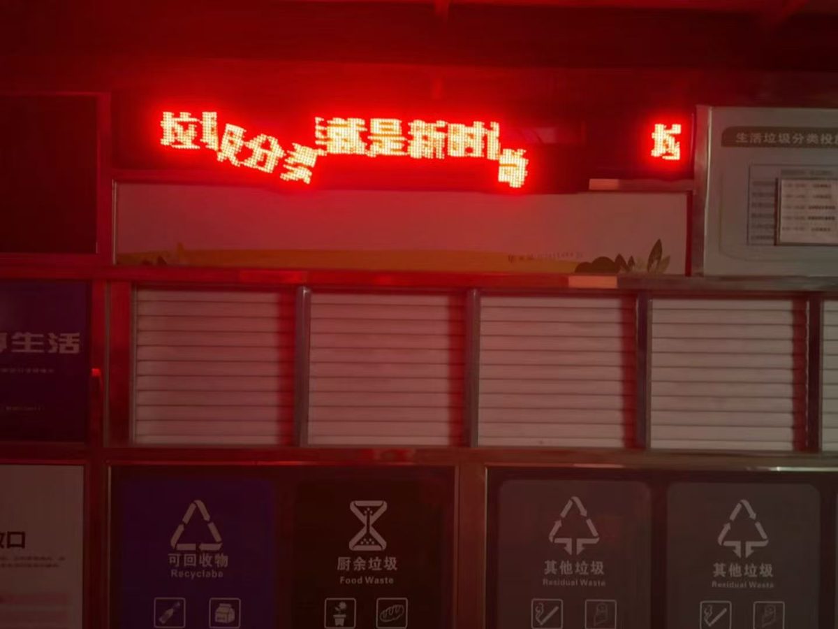 A community waste-sorting station. The LED display panels above the bins have slipped out of alignment, so the red characters of the slogan float at different heights.